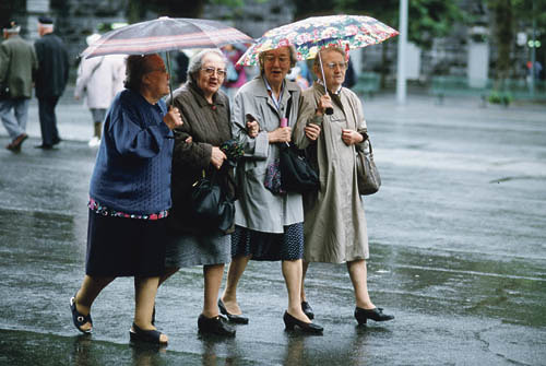 Vier Seniorinnen spazieren mit Regenschirmen bei Regenwetter Arm in Arm auf der Stra?e