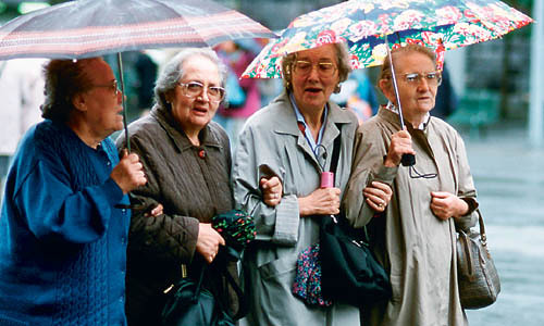 Vier Seniorinnen spazieren mit Regenschirmen bei Regenwetter Arm in Arm auf der Stra?e